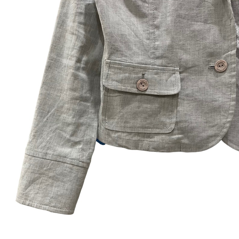 Puede incluir: Una blazer gris claro con un corte entallado. La chaqueta presenta solapas con muescas, dos bolsillos delanteros y botones con un logotipo. Las mangas tienen un detalle de pu&ntilde;o abotonado. La blazer est&aacute; hecha de una tela texturizada.