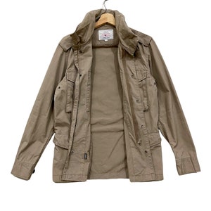 K&ouml;nnte beinhalten: Eine beige Jacke mit mehreren Taschen, einem hohen Kragen und einem Rei&szlig;verschluss. Die Jacke h&auml;ngt an einem Holzb&uuml;gel. Ein kleines Etikett ist im Brustbereich sichtbar. Die Jacke scheint aus einem strapazierf&auml;higen Stoff zu sein.