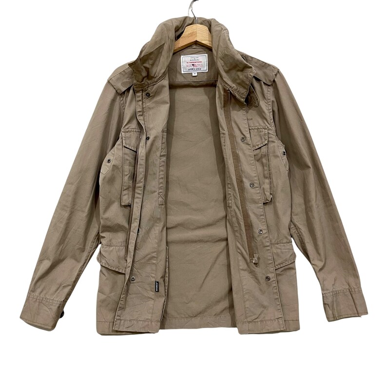 K&ouml;nnte beinhalten: Eine beige Jacke mit mehreren Taschen, einem hohen Kragen und einem Rei&szlig;verschluss. Die Jacke h&auml;ngt an einem Holzb&uuml;gel. Ein kleines Etikett ist im Brustbereich sichtbar. Die Jacke scheint aus einem strapazierf&auml;higen Stoff zu sein.