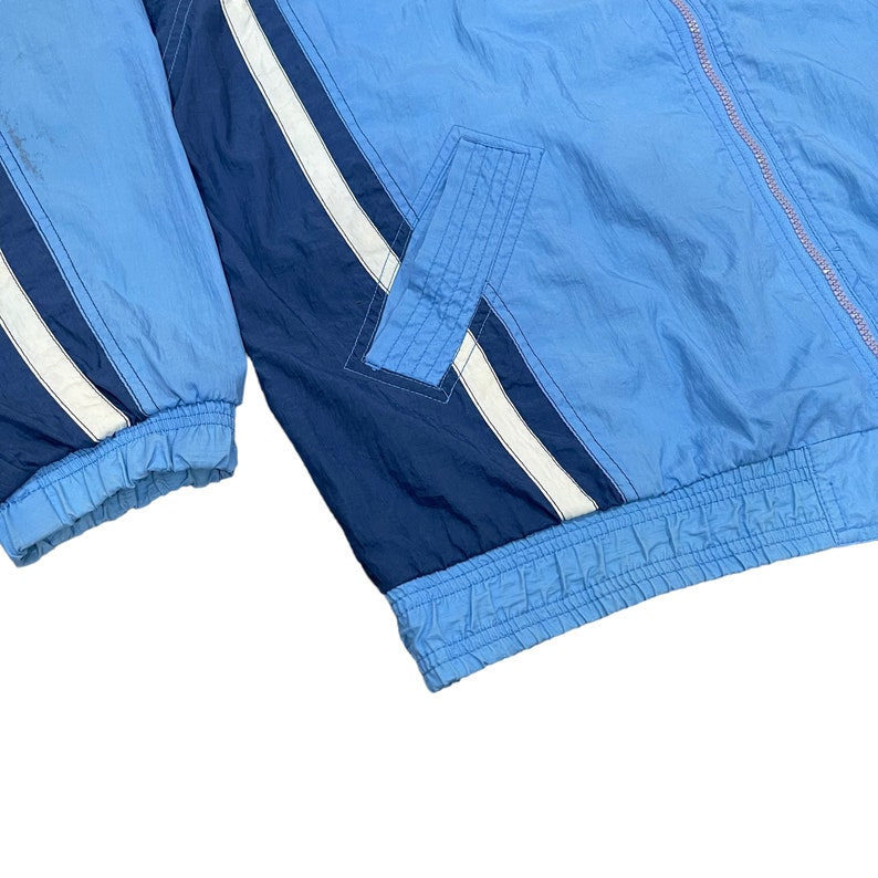 May include: A light blue and navy blue windbreaker jacket with white stripes. The jacket has a front pocket and a zipper closure. The cuffs and hem are elasticized. The jacket is made of a lightweight, possibly nylon, material.