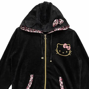 Pode incluir: Casaco de moletom preto de veludo com z&iacute;per e forro de estampa de leopardo rosa. O moletom tem um design Hello Kitty na frente e um la&ccedil;o rosa no capuz.