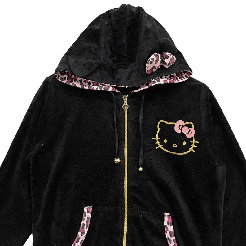 Pode incluir: Casaco de moletom preto de veludo com z&iacute;per e forro de estampa de leopardo rosa. O moletom tem um design Hello Kitty na frente e um la&ccedil;o rosa no capuz.