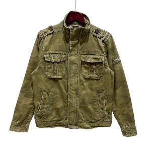Op de afbeelding: Een groene camouflagejas met een ritssluiting en twee klepzakken aan de voorkant. De jas heeft een hoge kraag en een merklabel op de rechtermouw met de tekst "Abercrombie & Fitch".