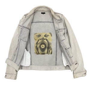 May include: A light gray denim jacket with a graphic print of a face on the back. The jacket is unbuttoned and the lining is visible.