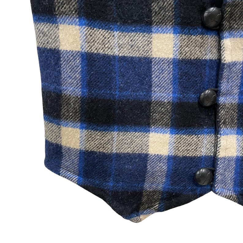 May include: A vest featuring a plaid pattern in shades of blue, black, and beige. The garment has a textured fabric surface and is fastened along the right side with three round, black, textured buttons arranged in a vertical line.