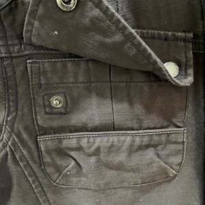 K&ouml;nnte beinhalten: Ein dunkelbraunes Kleidungsst&uuml;ck mit einer rechteckigen aufgesetzten Tasche, deren Klappe mit einem metallischen Druckknopf verschlossen wird. Die Taschenklappe wird ebenfalls durch einen passenden Metalldruckknopf gehalten, und der Stoff zeigt eine sichtbare Webstruktur.