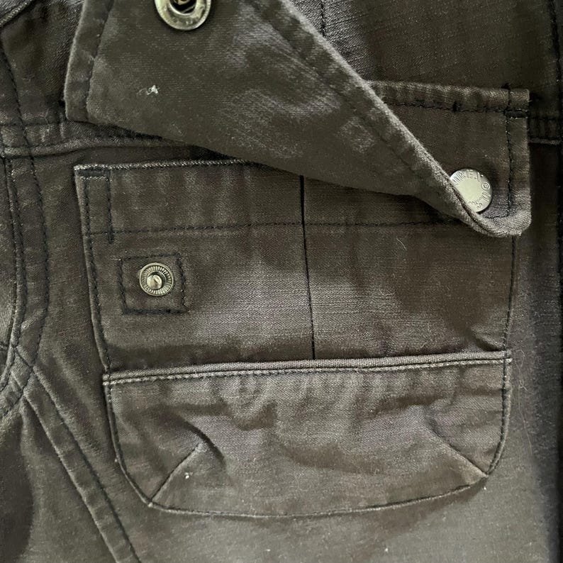 K&ouml;nnte beinhalten: Ein dunkelbraunes Kleidungsst&uuml;ck mit einer rechteckigen aufgesetzten Tasche, deren Klappe mit einem metallischen Druckknopf verschlossen wird. Die Taschenklappe wird ebenfalls durch einen passenden Metalldruckknopf gehalten, und der Stoff zeigt eine sichtbare Webstruktur.