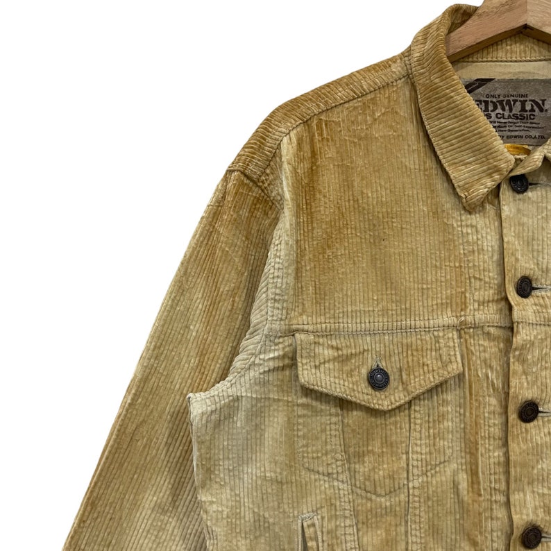 May include: A close-up shot of a tan corduroy jacket with a button-down front and two chest pockets. The jacket features a classic collar and is made of ribbed fabric. The brand name "EDWIN" is visible on a tag.