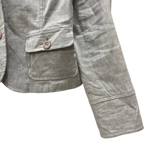 Puede incluir: Un blazer gris claro con dos bolsillos delanteros y un cierre de bot&oacute;n &uacute;nico. El blazer tiene un corte entallado y un dise&ntilde;o cl&aacute;sico. La manga es visible, mostrando un detalle de pu&ntilde;o. La tela tiene una textura sutil.