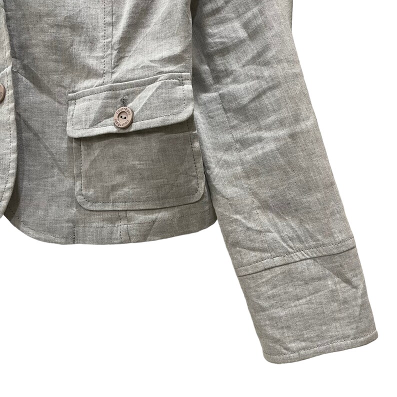 Puede incluir: Un blazer gris claro con dos bolsillos delanteros y un cierre de bot&oacute;n &uacute;nico. El blazer tiene un corte entallado y un dise&ntilde;o cl&aacute;sico. La manga es visible, mostrando un detalle de pu&ntilde;o. La tela tiene una textura sutil.