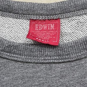 May include: Close-up of a gray sweatshirt with a red tag. The tag reads "EDWIN" in white capital letters. Below the brand name is smaller text. The tag also has the word "SMALL" printed on it.
