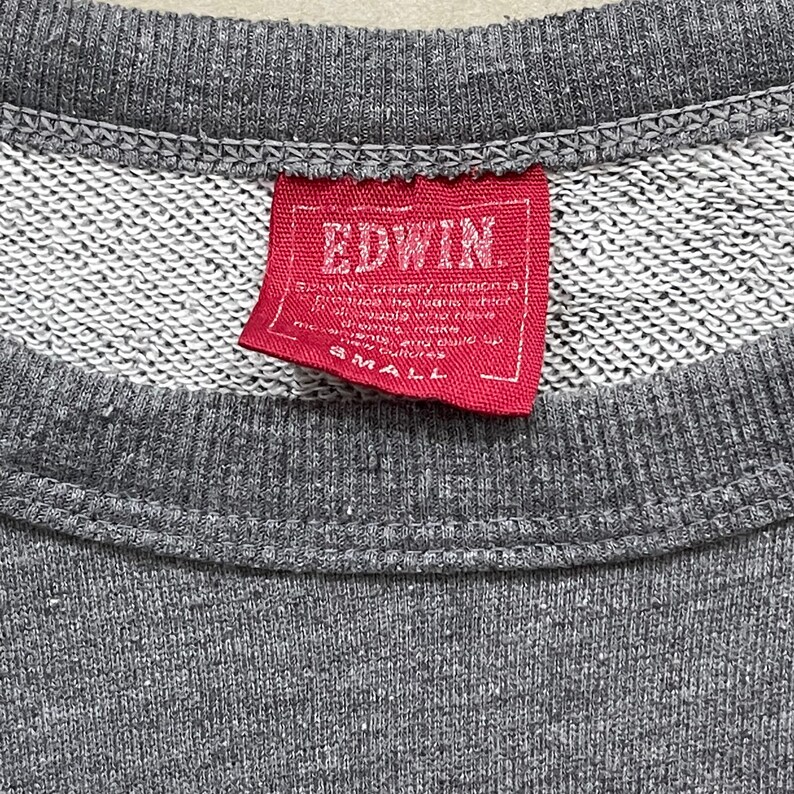 May include: Close-up of a gray sweatshirt with a red tag. The tag reads "EDWIN" in white capital letters. Below the brand name is smaller text. The tag also has the word "SMALL" printed on it.