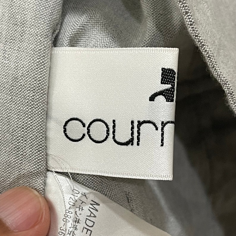 Puede incluir: Primer plano de una etiqueta de ropa blanca con el nombre de la marca "courr" en negro, fuente sans-serif. Un gr&aacute;fico negro est&aacute; encima del nombre de la marca. La etiqueta est&aacute; cosida en una tela gris. La etiqueta tambi&eacute;n tiene texto japon&eacute;s y un n&uacute;mero de tel&eacute;fono.
