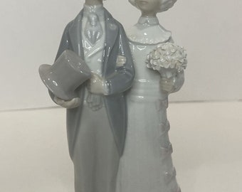 May include: A porcelain figurine of a bride and groom. The bride is wearing a white dress with a floral pattern and a white veil. The groom is wearing a gray suit with a top hat. The figurine is in a traditional wedding pose.