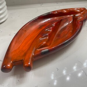 May include: A ceramic ashtray shaped like a leaf with a glossy orange glaze. The ashtray has three indentations for cigarettes.