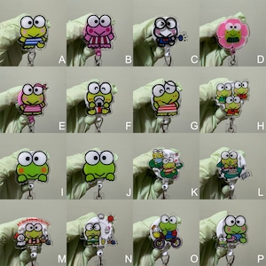 Op de afbeelding: Een set van 16 verschillende Keroppi-personage-badge-rollen. Elke rol heeft een ander Keroppi-ontwerp, waaronder Keroppi in een badpak, Keroppi met een drankje, Keroppi met vrienden en Keroppi op een fiets. De rollen zijn gemaakt van helder plastic en hebben een metalen clip.