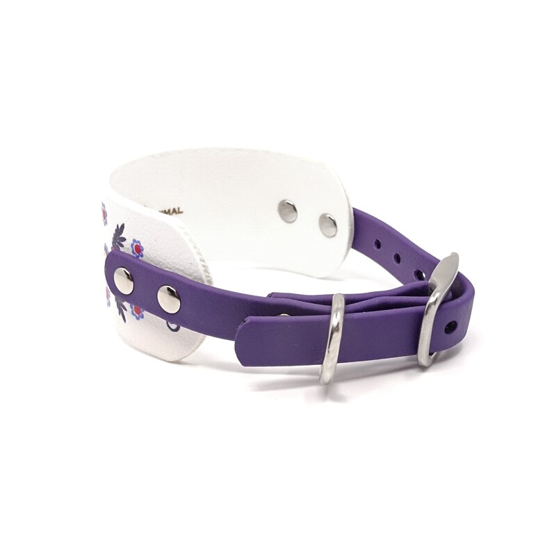 Puede incluir: Collar de perro de cuero blanco con correa morada y hebilla plateada. El collar tiene un dise&ntilde;o floral con flores rojas, azules y blancas.