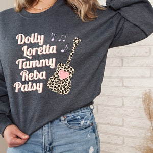 May include: A dark gray sweatshirt with a pink and black leopard print guitar design. The guitar has a pink heart on it. The text "Dolly Loretta Tammy Reba Patsy" is printed on the front of the sweatshirt.