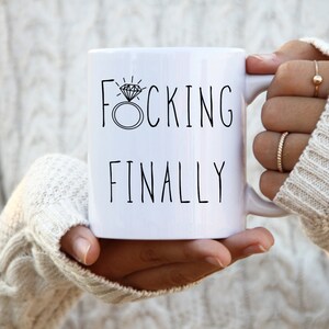May include: White ceramic coffee mug with black text that reads "F*CKING FINALLY" with a diamond ring illustration.