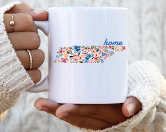 Puede incluir: Taza de cerámica blanca con un diseño floral del estado de Tennessee y la palabra "home" en letras azules.