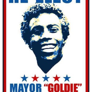 Può includere: Un manifesto di campagna politica con il ritratto di un uomo sorridente. Il manifesto recita "RE-ELECT MAYOR 'GOLDIE' WILSON" in testo blu e rosso, con cinque stelle sotto il ritratto. Il manifesto ha un bordo rosso e blu.