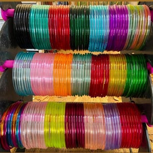 May include: A collection of translucent, circular bangles arranged in rows on a black display stand. The bangles feature a wide spectrum of colours including black, teal, red, dark green, blue, purple, pink, orange, yellow, and magenta.
