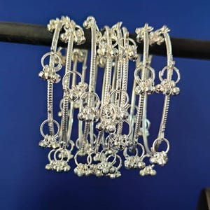 May include: A set of silver bangles with detailed floral and bell-shaped embellishments. The bangles are presented against a dark blue backdrop, highlighting their delicate design and reflective quality. Each bangle has a diameter of approximately 6.35 cm.