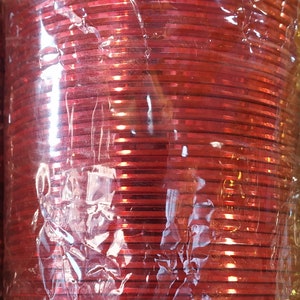 May include: A stack of red glass bangles wrapped in plastic. The bangles are shiny and have a metallic finish.