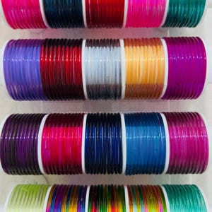 Puede incluir: Una colección de pulseras de vidrio coloridas en varios tonos, incluyendo rosa, rojo, azul y verde. Las pulseras están dispuestas en filas, mostrando un espectro de colores. Cada conjunto se mantiene unido por una base cilíndrica blanca.