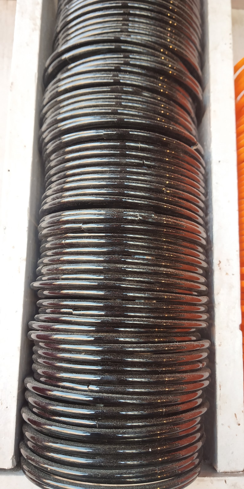 May include: A stack of black bangles, probably made of glass or plastic, arranged in a vertical column. The bangles are shiny and have a slight sheen.