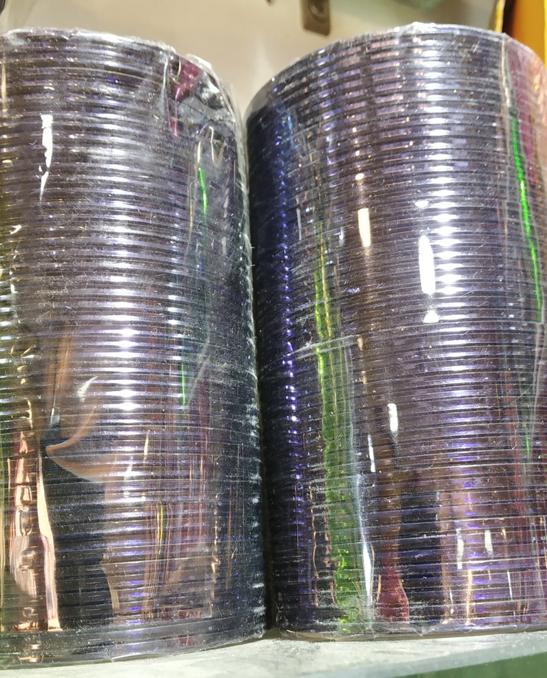 May include: Two stacks of iridescent purple bangles wrapped in clear plastic. The bangles are stacked on top of each other and are ready to be sold.