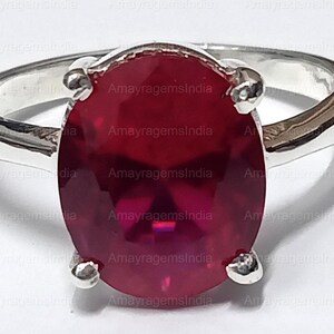 May include: A silver ring with a large, oval-shaped, red gemstone. The gemstone is set in a silver prong setting. The ring is on a white background.