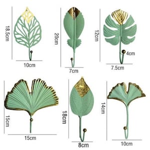 Puede incluir: Conjunto de seis ganchos de pared decorativos con forma de hoja, en tonos verdes y dorados. Las medidas están en centímetros. Los diseños incluyen varias formas de hojas, cada uno con un gancho para colgar objetos.