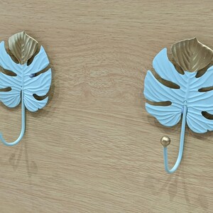 Puede incluir: Dos ganchos decorativos de pared con forma de hojas de monstera. Las hojas son de color azul claro con detalles dorados y un gancho curvo. Montados sobre una superficie de madera clara.