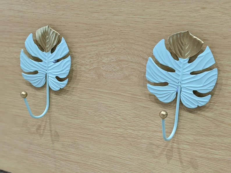 Puede incluir: Dos ganchos decorativos de pared con forma de hojas de monstera. Las hojas son de color azul claro con detalles dorados y un gancho curvo. Montados sobre una superficie de madera clara.