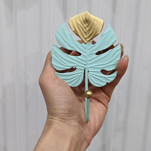 Puede incluir: Un gancho de pared decorativo con forma de hoja de monstera. La hoja es de color azul claro con detalles dorados. El gancho tiene una bola dorada en el extremo. El gancho es de metal.