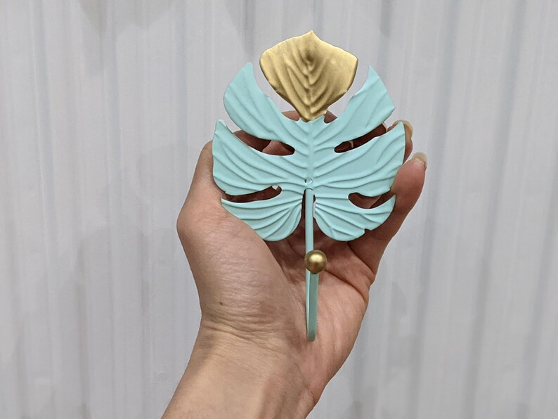 Puede incluir: Un gancho de pared decorativo con forma de hoja de monstera. La hoja es de color azul claro con detalles dorados. El gancho tiene una bola dorada en el extremo. El gancho es de metal.