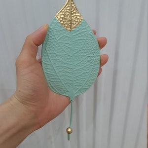 Puede incluir: Un colgador de pared decorativo con forma de hoja, de color verde suave con una superficie texturizada. La parte superior es dorada y el gancho tiene una peque&ntilde;a bola dorada en el extremo. Ideal para decoraci&oacute;n inspirada en la naturaleza.