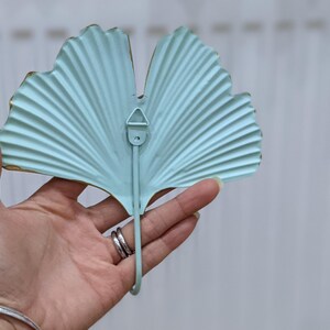 Puede incluir: Un colgador de pared de metal azul claro con forma de hoja de ginkgo. El gancho tiene un dise&ntilde;o curvo y un peque&ntilde;o lazo para colgar. Los bordes est&aacute;n adornados con oro. El gancho se sostiene en una mano.