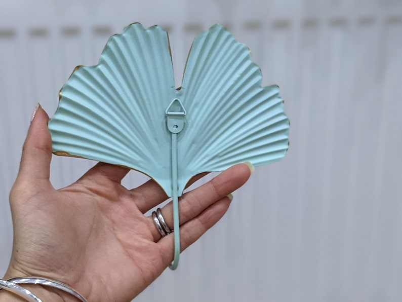 Puede incluir: Un colgador de pared de metal azul claro con forma de hoja de ginkgo. El gancho tiene un dise&ntilde;o curvo y un peque&ntilde;o lazo para colgar. Los bordes est&aacute;n adornados con oro. El gancho se sostiene en una mano.