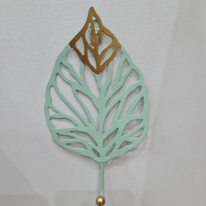 Puede incluir: Un gancho decorativo de pared con forma de hoja. La hoja es de color verde claro con una secci&oacute;n superior dorada. El gancho es de color verde claro con una bola dorada en el extremo.