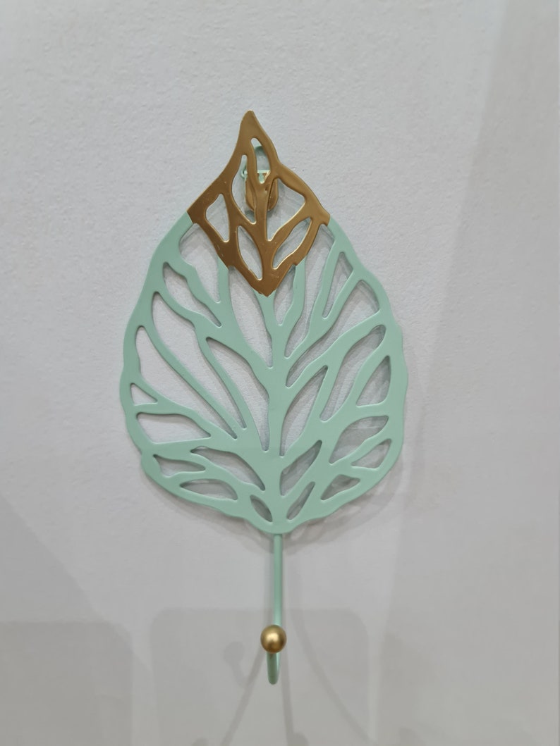 Puede incluir: Un gancho decorativo de pared con forma de hoja. La hoja es de color verde claro con una secci&oacute;n superior dorada. El gancho es de color verde claro con una bola dorada en el extremo.