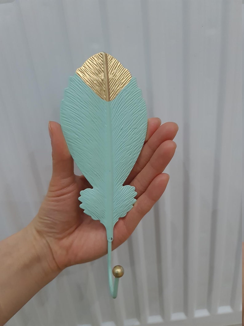 Puede incluir: Un gancho de pared decorativo con forma de pluma. La parte superior de la pluma es dorada y el resto es de color verde azulado claro. El gancho tiene una peque&ntilde;a bola dorada en el extremo.