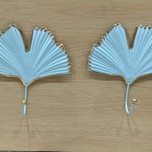 Puede incluir: Dos perchas de pared en forma de hoja azul claro con ribete dorado y un gancho dorado. Los ganchos est&aacute;n montados sobre una superficie de madera marr&oacute;n claro. El dise&ntilde;o de la hoja tiene una textura acanalada.