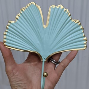 Puede incluir: Un gancho de pared decorativo con forma de hoja de ginkgo. La hoja es de color verde azulado claro con ribetes dorados. El gancho es del mismo color con una bola dorada en el extremo.