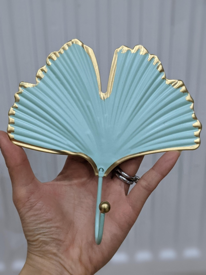 Puede incluir: Un gancho de pared decorativo con forma de hoja de ginkgo. La hoja es de color verde azulado claro con ribetes dorados. El gancho es del mismo color con una bola dorada en el extremo.