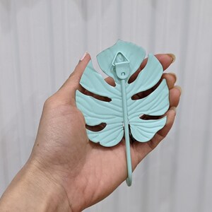 Puede incluir: Un gancho de pared de metal azul claro con forma de hoja de monstera. El gancho tiene un dise&ntilde;o de hoja y un gancho curvo. La parte posterior tiene un peque&ntilde;o lazo de metal para colgar.