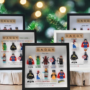 May include: Several framed artworks featuring the word "DADDY" in wooden tiles, with Lego superhero figures. The text "You are..." is above the figures, with additional text below. The frames are black and white, set against a blurred background.