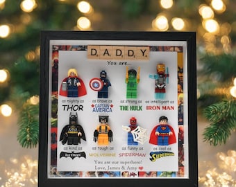 May include: A framed artwork featuring Lego superhero figures, including Thor, Captain America, and Batman. The text reads "DADDY, You are..." with personalized messages below. The frame is black, and the background is a festive scene.