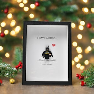 Può includere: Un'opera d'arte incorniciata in nero con un personaggio Lego Batman e il testo "I HAVE A HERO... I CALL HIM HASAN LOVE, HENRY" e un cuore rosso. Lo sfondo è bianco.